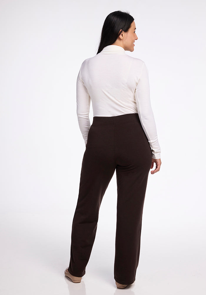 A person with long dark hair is standing, facing away. They are wearing a white long-sleeve top and Woolx's Ellie Wide Leg Pants in French Roast, which feature subtle side pockets. The background is plain and light-colored.