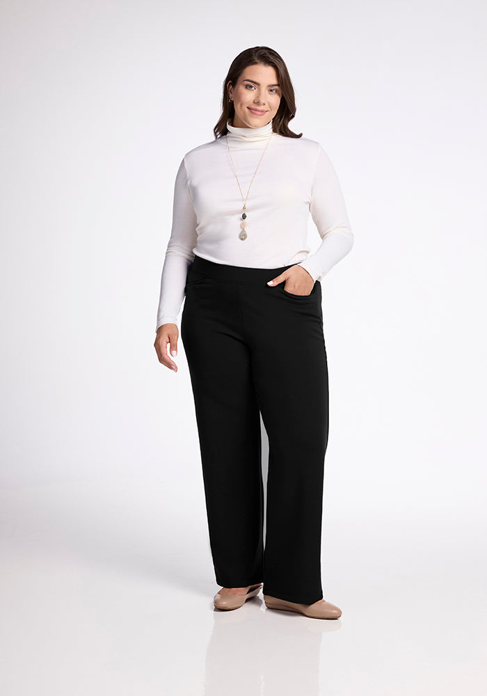 A woman stands against a plain white background, wearing a white turtleneck top and the Ellie Pants Tall by Woolx, featuring an elastic waistband. She has one hand in her deep front pocket and is smiling while accessorizing with a long necklace and beige flats.