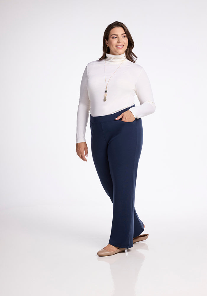 A woman stands confidently, wearing a white turtleneck and Woolx's Ellie Wide Leg Pants in Deep Navy, paired with beige flats. She has a long necklace and her hand in her pant pocket, smiling against a plain white background.