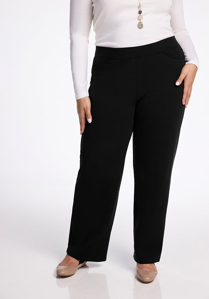 A person is wearing a long-sleeved white top and high-waisted black Ellie Wide Leg Pants Tall by Woolx, standing with one hand on their hip. They are also wearing nude-colored shoes and a necklace with round pendants against a plain white background. 