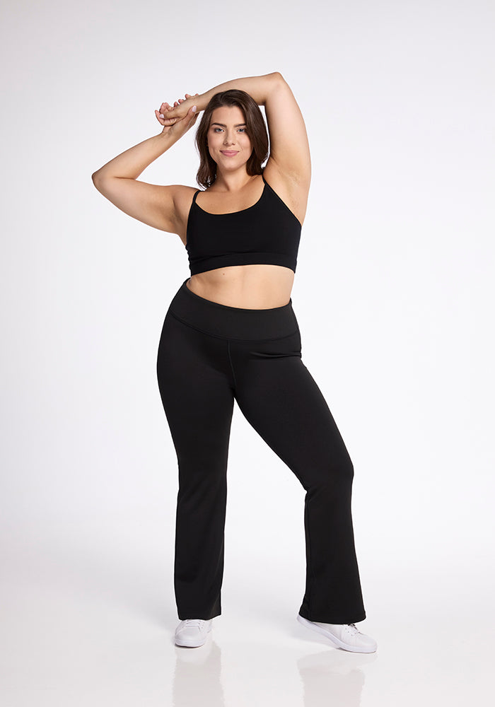 A woman stands confidently against a plain white background. She is wearing a black crop top and Woolx's Marissa Pants in black, paired with white sneakers. One of her arms is raised and slightly bent above her head, while the other rests at her side. She smiles gently at the camera.