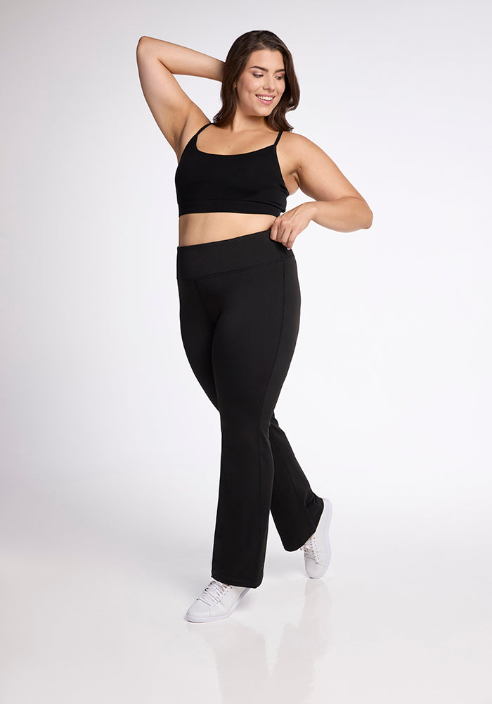 A woman wearing a black sports bra and the Woolx Marissa Pants in black poses confidently against a plain white background. She is smiling with one arm bent over her head and the other hand on her hip. She is also wearing white sneakers.