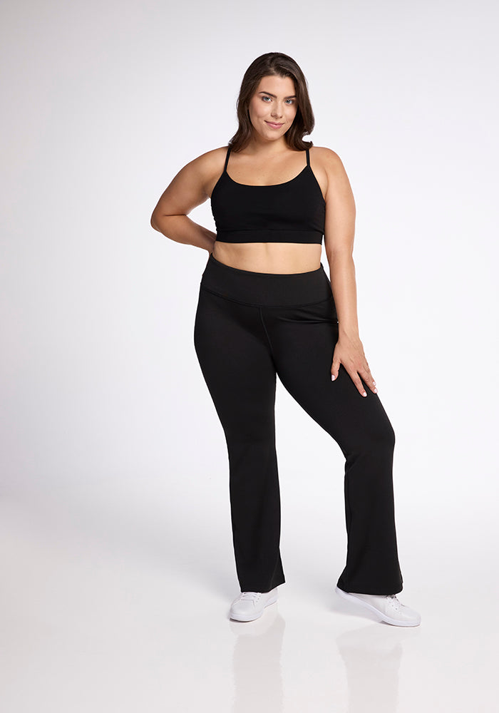 A woman stands confidently against a white background, wearing a black sports bra and Marissa Pants - Black by Woolx. She has one hand on her hip and the other resting on her thigh. She is also wearing white sneakers and has a slight smile on her face.