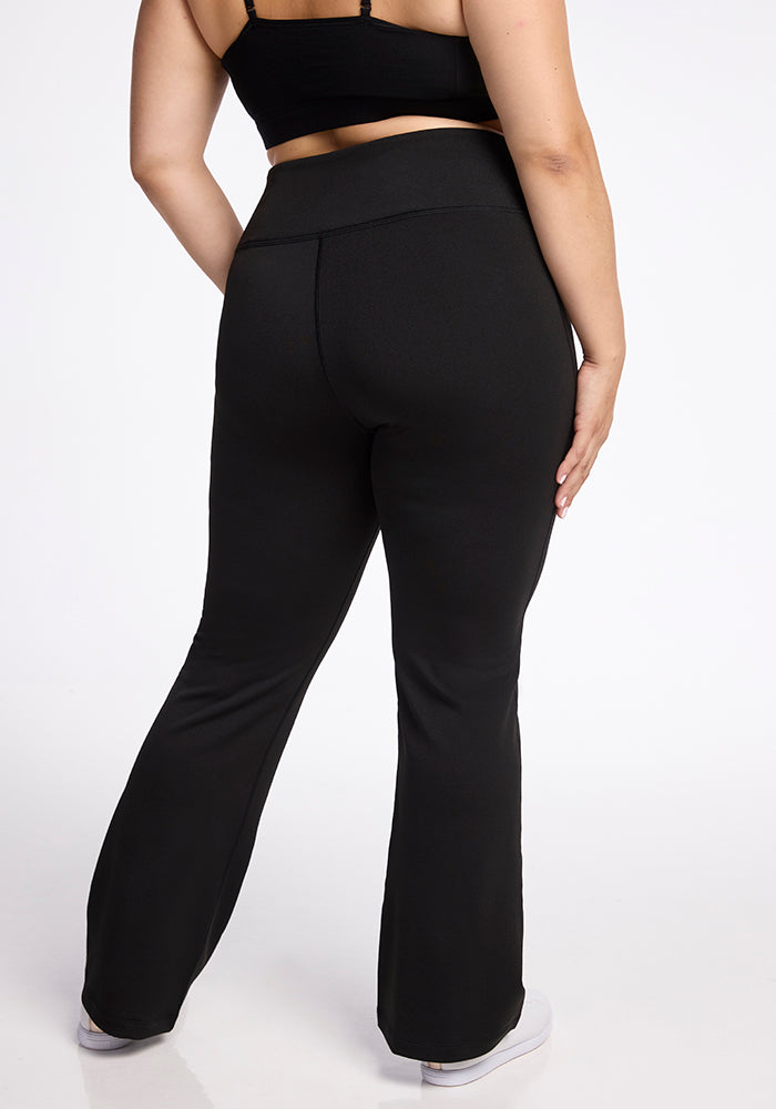 A person wearing the Marissa Pants - Black by Woolx and a black crop top is shown from behind. The individual stands against a plain white background, with their left hand resting on their hip and their right arm hanging by their side. They are also wearing white sneakers.