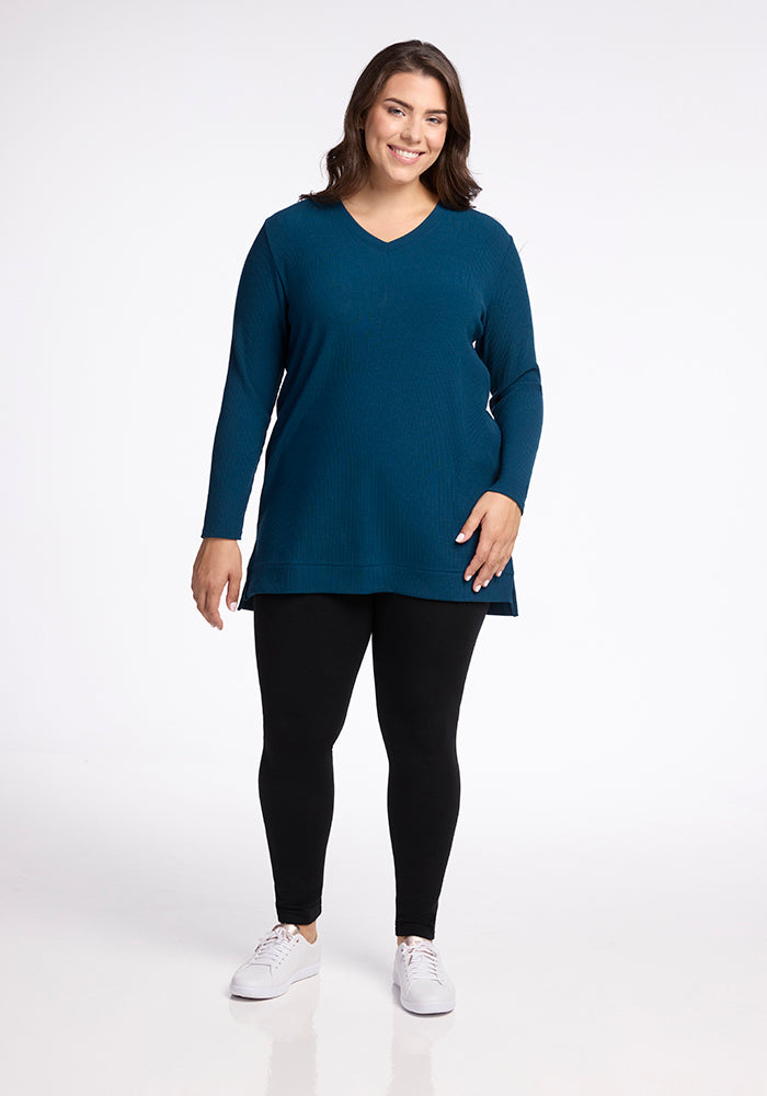 A smiling woman stands against a plain white background. She is wearing a cozy and warm Evie Tunic in Real Teal from Woolx, paired with black leggings and white sneakers. She has long, dark hair and is posing with one hand resting on her hip.