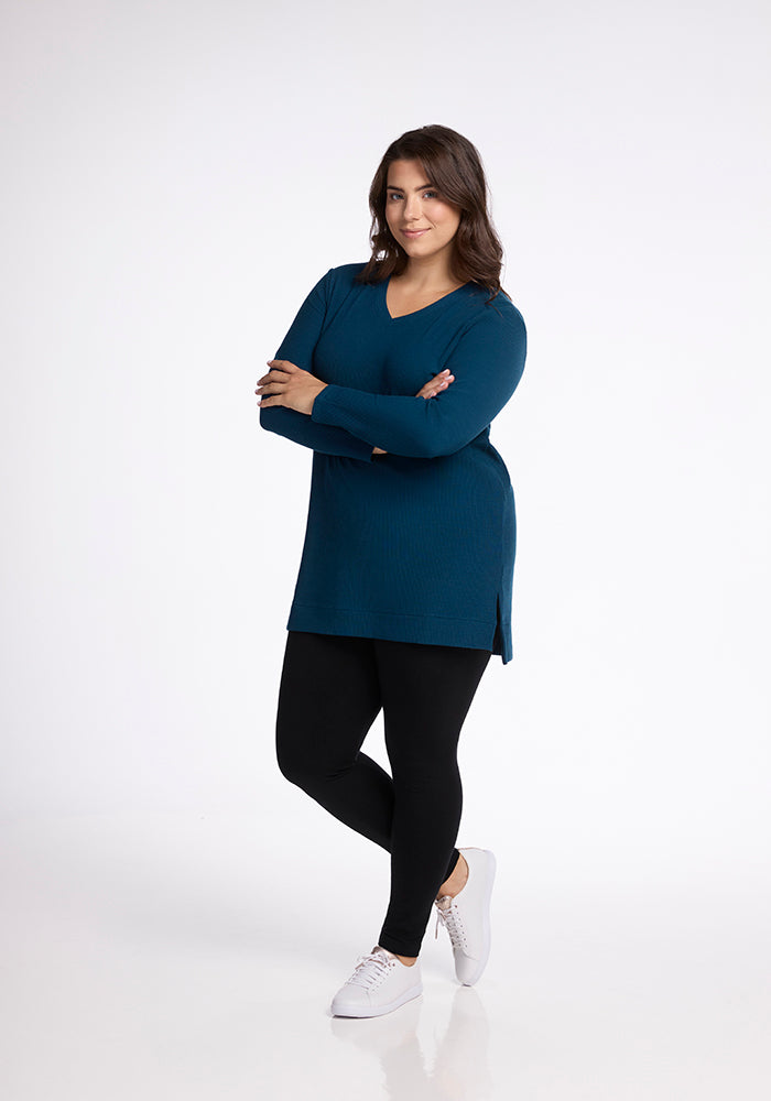 A person with long dark hair stands with arms crossed, dressed in the Woolx "Evie Tunic" in Real Teal, black leggings, and white sneakers. They are smiling and standing against a plain white background.