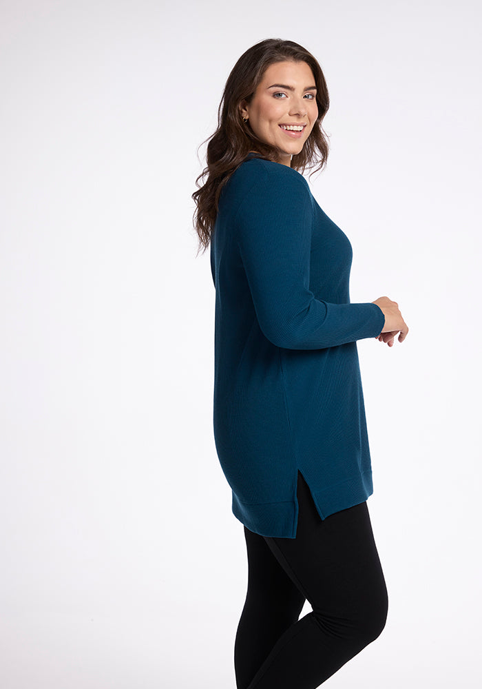 A woman with long brown hair smiles while looking over her shoulder. She is wearing a cozy, teal Evie Tunic from Woolx paired with black leggings, offering a flattering fit. The background is plain and white.