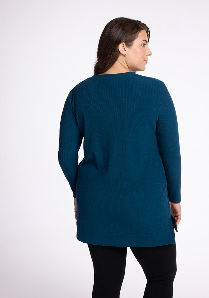 A woman stands facing away from the camera, wearing an Evie Tunic - Real Teal by Woolx, made of cozy Merino wool, paired with black leggings. The flattering fit of the tunic complements her figure against the plain white background.