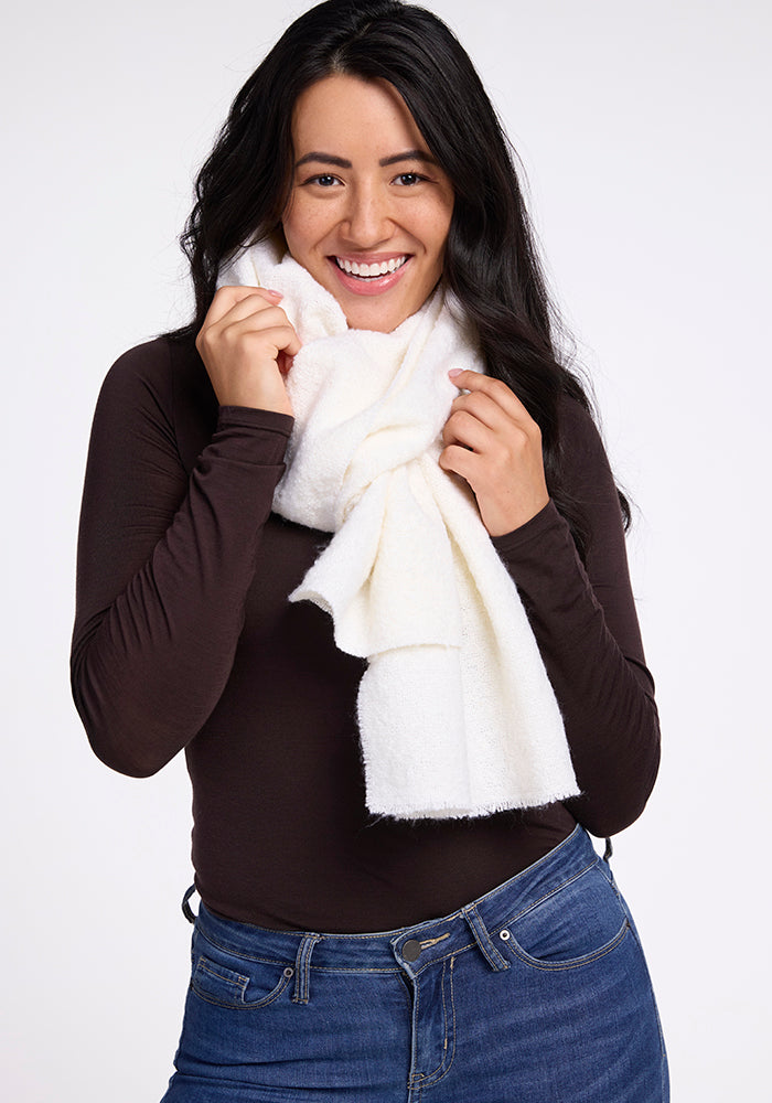 A person with long dark hair, wearing a black long-sleeve shirt and blue jeans, smiles while holding the Woolx Bradie Scarf in cream wrapped around their neck. The background is plain and light-colored.