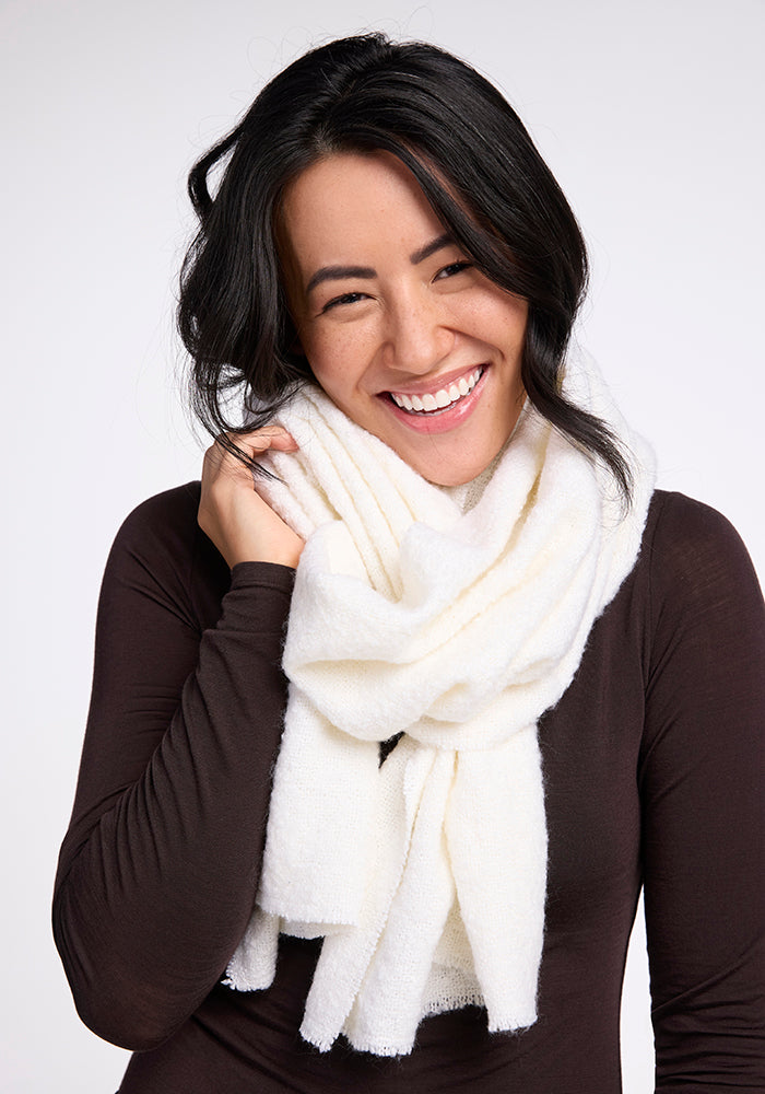 A woman with long dark hair is smiling and looking at the camera. She is wearing a dark long-sleeve top and has a thick, cozy Bradie Scarf - Cream from Woolx wrapped around her neck. The background is plain white.