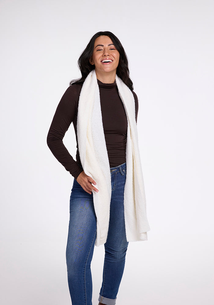 A woman with long dark hair is smiling and standing against a plain background. She is wearing a dark long-sleeve top, blue jeans, and the Bradie Scarf in cream from Woolx draped around her neck.