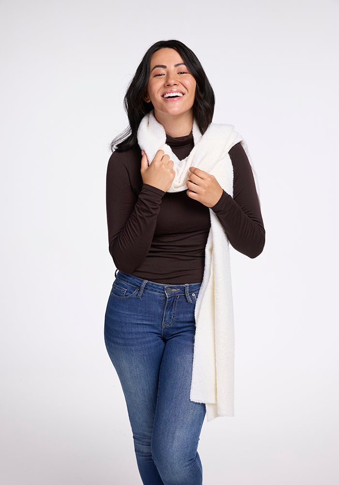 A person with long dark hair is smiling while wearing a dark long-sleeve top, jeans, and the Bradie Scarf in cream from Woolx. They have one hand touching the scarf and the other hand resting by their side. The background is plain and white.