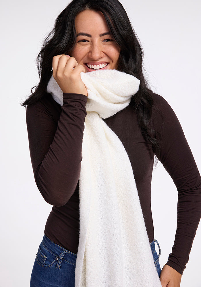 A woman with long dark hair, wearing a black long-sleeve top and blue jeans, smiles brightly while holding the Woolx Bradie Scarf in Cream close to her face and around her neck. The background is plain and light-colored.