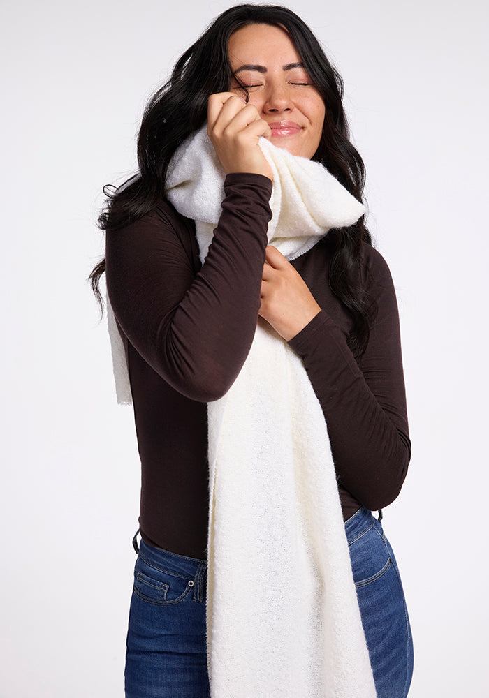 A woman with long dark hair wearing a black long-sleeve top and blue jeans hugs the Woolx Bradie Scarf in Cream to her face with a content and happy expression. The background is plain white.