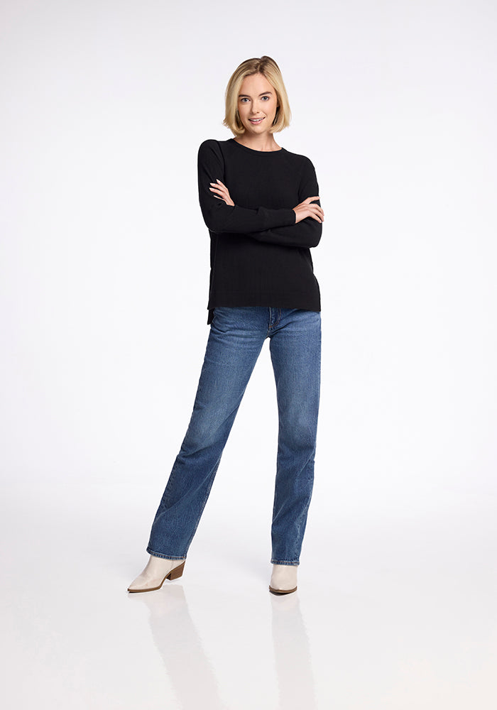 A woman with shoulder-length blonde hair stands confidently with arms crossed. She is wearing the Ainsley Sweater in black by Woolx, paired with blue jeans and white ankle boots. The background is plain white.