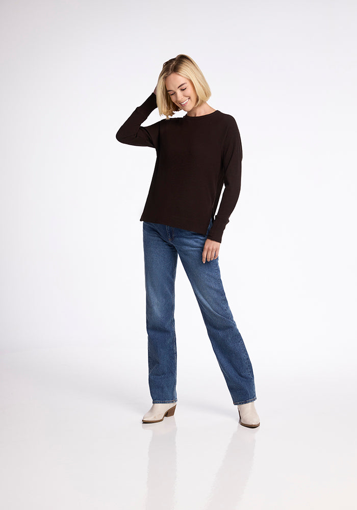 A person with short blonde hair stands against a plain backdrop, smiling and looking down. They are wearing an Ainsley Sweater in French Roast by Woolx, blue jeans, and white shoes. Their left hand is touching their head, and their right hand is relaxed by their side, showcasing a versatile style.