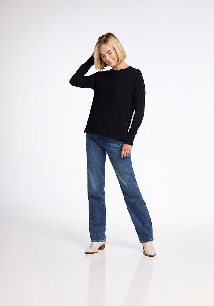 A person with short blonde hair stands against a plain background, smiling and looking down. They are wearing a long-sleeve Ainsley Sweater - Black from Woolx, blue jeans, and light-colored shoes. One hand is raised to their head, and the other rests by their side.