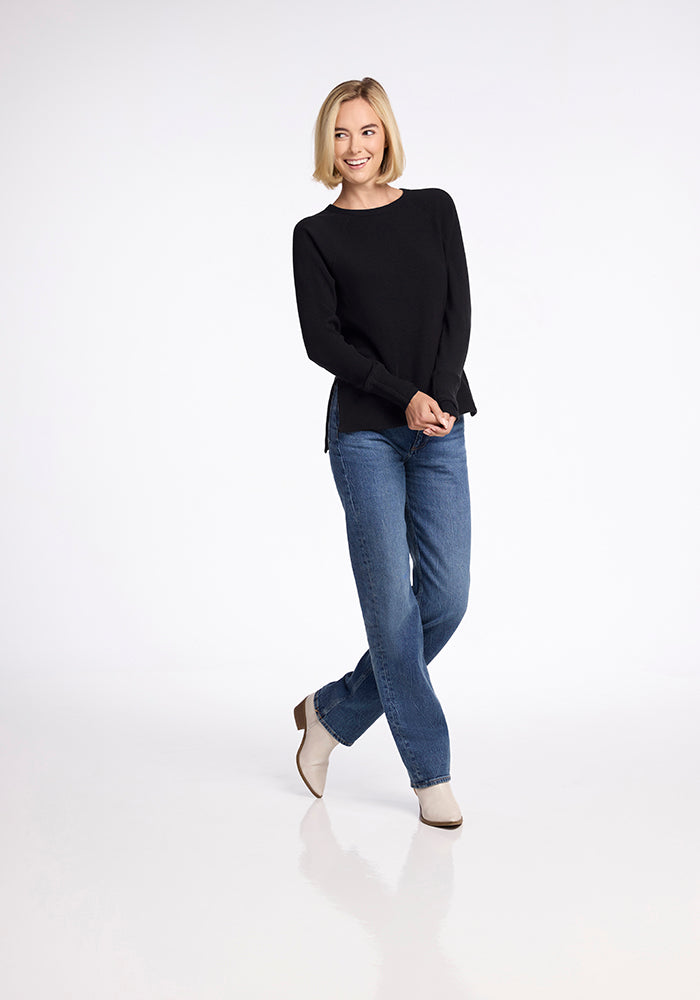 A person with short, light-colored hair stands against a plain background. They are wearing the Ainsley Sweater in black by Woolx, along with blue jeans and beige boots. They are smiling and standing with one leg slightly crossed in front of the other.