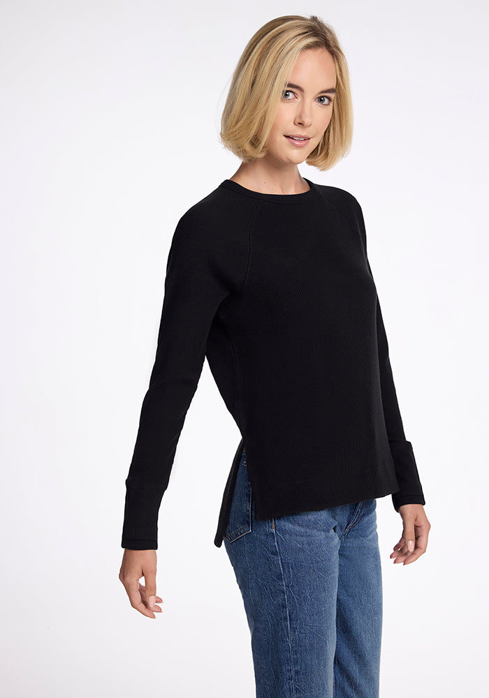 A person with short blonde hair is standing against a plain background, wearing the Ainsley Sweater in black from Woolx and blue jeans. The person is looking directly at the camera with a slight smile and a relaxed pose. 