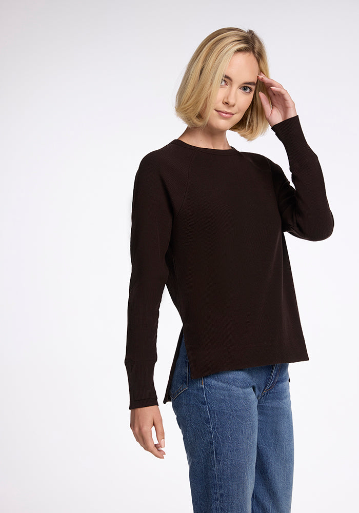 A person with short blonde hair is wearing a dark long-sleeve Ainsley Sweater in French Roast from Woolx and blue jeans. They are standing against a neutral background, facing slightly to the right while touching their head with their left hand, showcasing their versatile style.