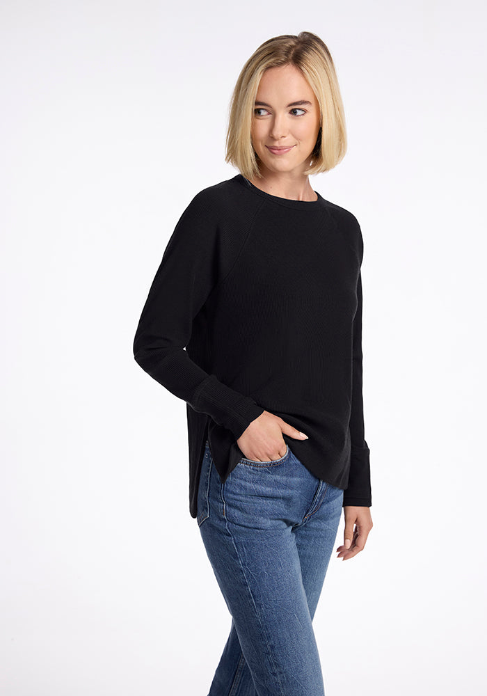 A person with shoulder-length blonde hair is standing against a plain white background. They are wearing the Woolx Ainsley Sweater in black and blue jeans, with one hand in their pocket. They are looking slightly to the side with a neutral expression.