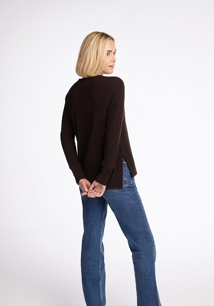 A person with shoulder-length blonde hair, wearing a dark brown Woolx Ainsley Sweater in the French Roast color and blue jeans, stands with their back to the camera and hands clasped behind them. The background is plain white.