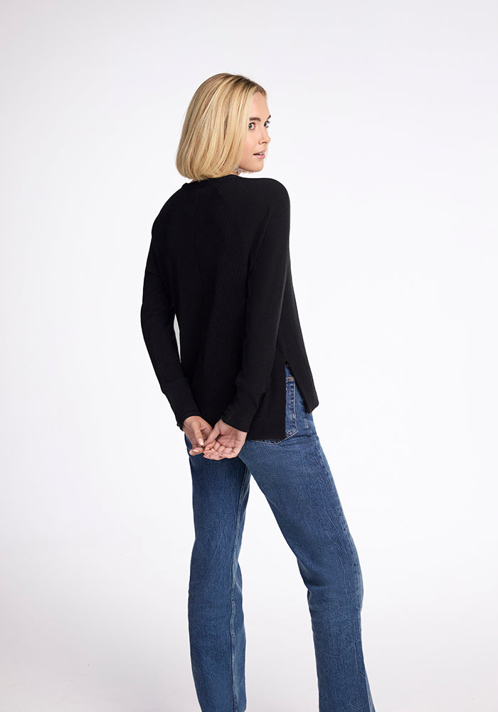 A person with blonde hair, wearing the Woolx Ainsley Sweater in black and blue jeans, stands against a white background. They are facing away, looking over their right shoulder with their hands clasped behind their back.