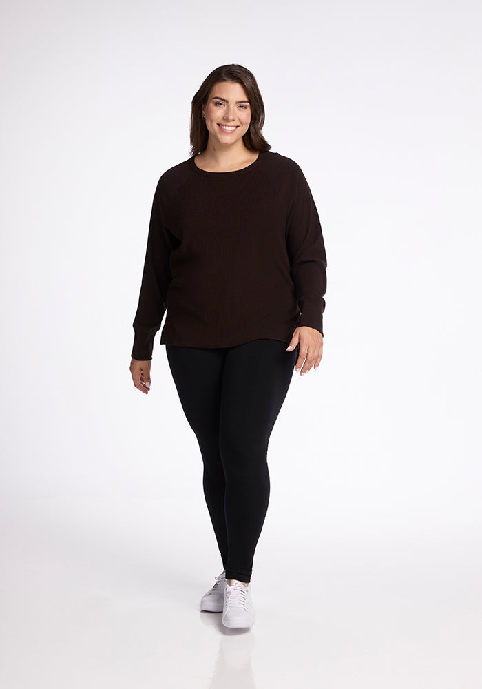 A smiling woman with medium-length brown hair is standing against a white background. She is wearing the Woolx Ainsley Sweater in French Roast, paired with black leggings and white sneakers. Her relaxed and confident posture showcases her versatile style effortlessly.