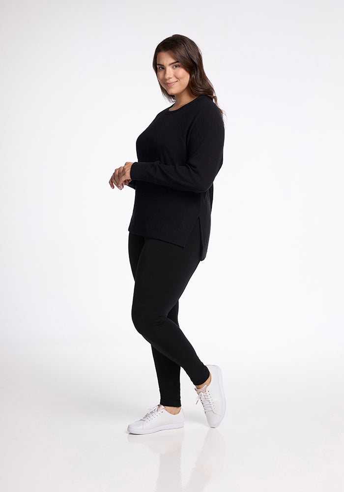 A woman stands against a plain background, smiling and wearing the Woolx Ainsley Sweater in black along with black leggings and white sneakers. She has long, wavy dark hair and is posed with her hands lightly clasped.