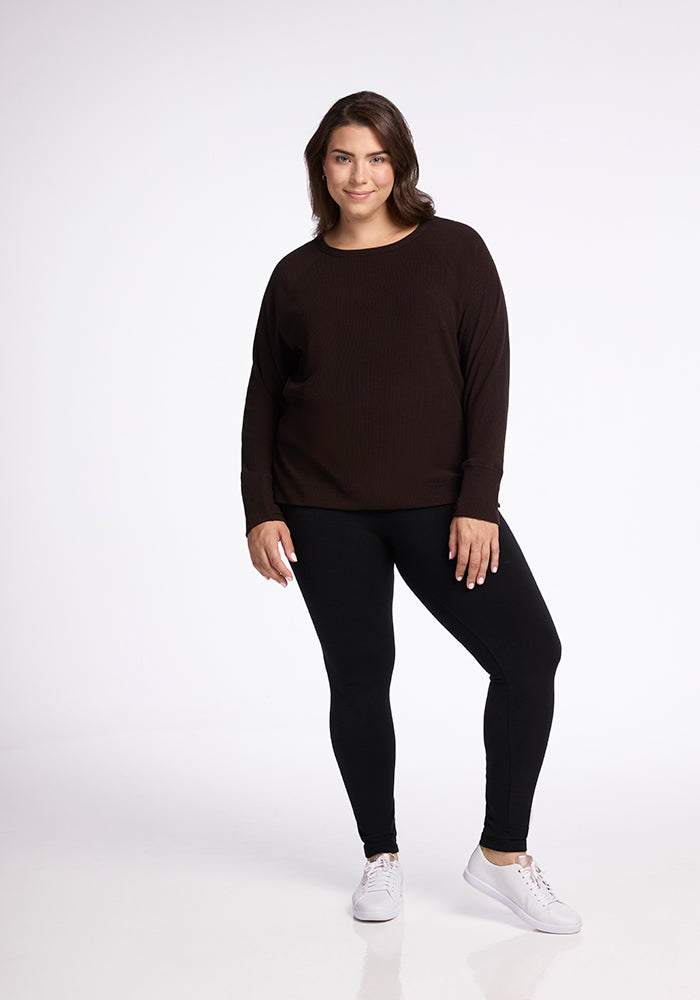 A person with long dark hair stands against a plain white background, wearing the versatile Ainsley Sweater in French Roast by Woolx, black leggings, and white sneakers. They are smiling and have one hand resting beside their hip while the other is relaxed by their side.