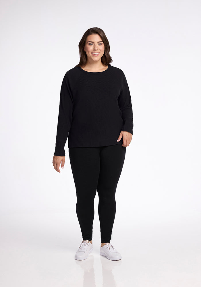 A woman is standing and smiling at the camera. She is wearing a Woolx Ainsley Sweater in black, paired with black leggings and white sneakers. The background is plain white.