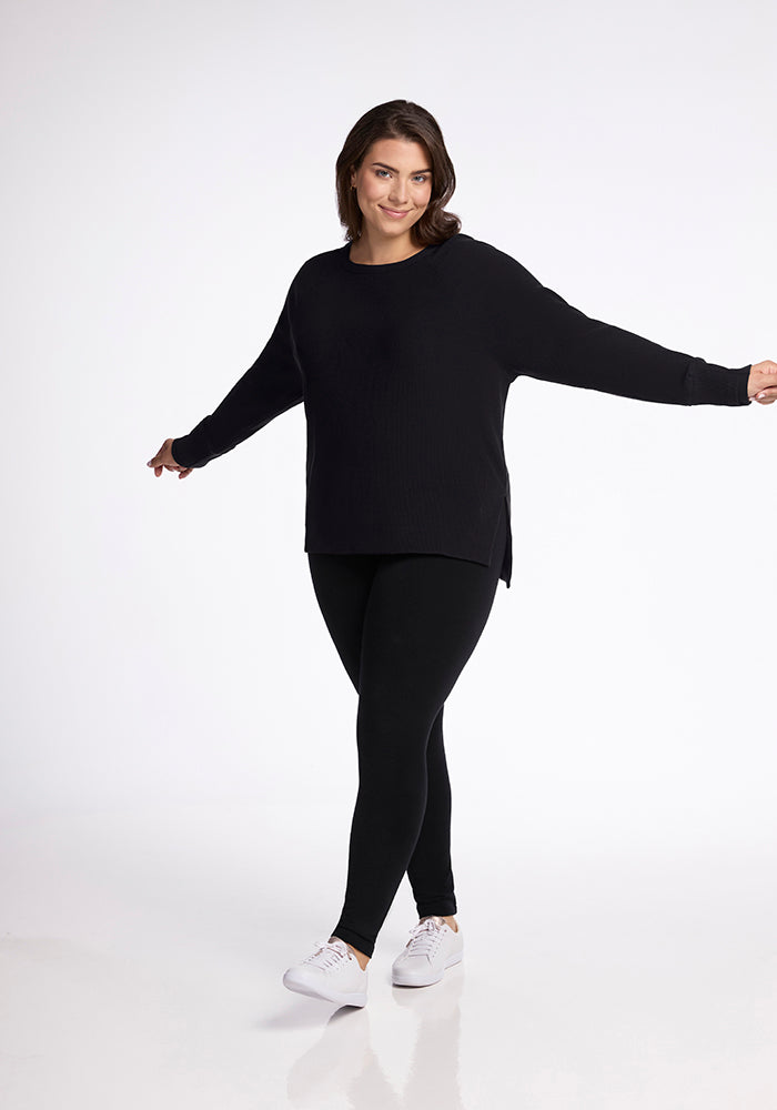 A person wearing a black Ainsley Sweater from Woolx and black leggings stands against a plain white background. They are smiling and extending their arms to the sides, and they are wearing white sneakers.