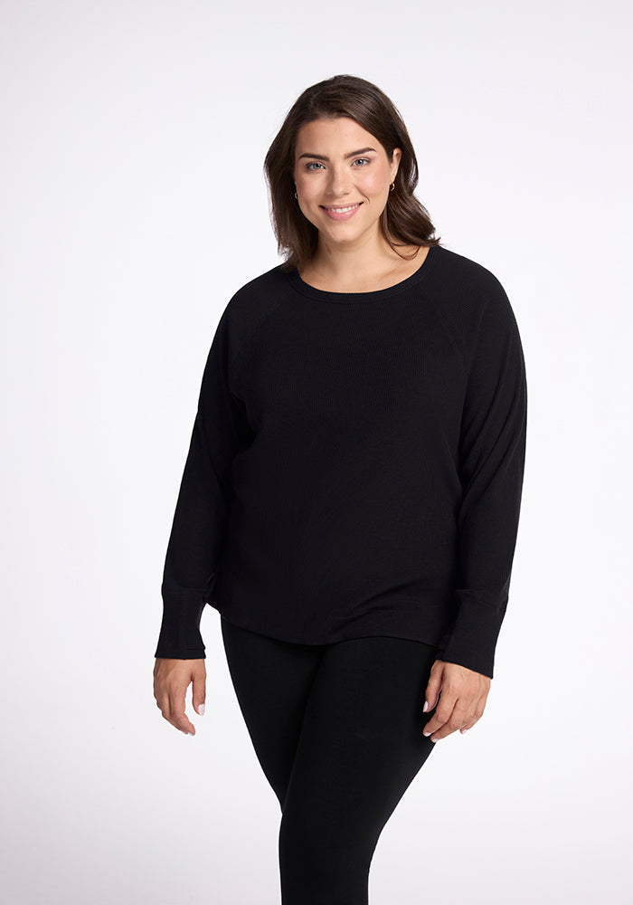 A woman with shoulder-length brown hair is smiling and wearing the Ainsley Sweater in black from Woolx, along with black pants. She stands against a plain white background. 