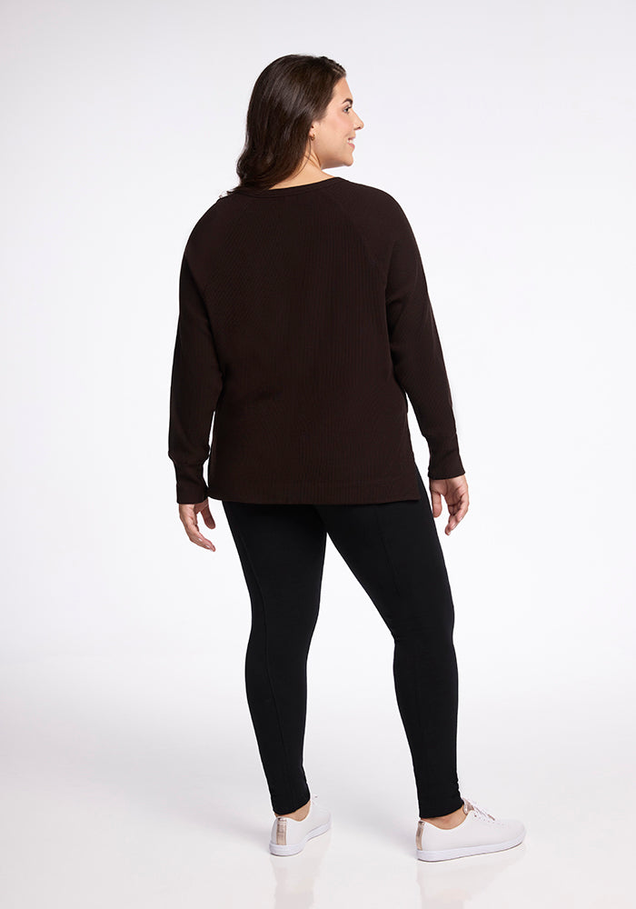 A woman dressed in a loose-fitting, long-sleeved Ainsley Sweater in French Roast by Woolx and black leggings is standing and looking to her right. With white sneakers on, she stands against a plain white background, facing away from the camera. The Merino Wool fabric adds to the versatile style of her outfit.