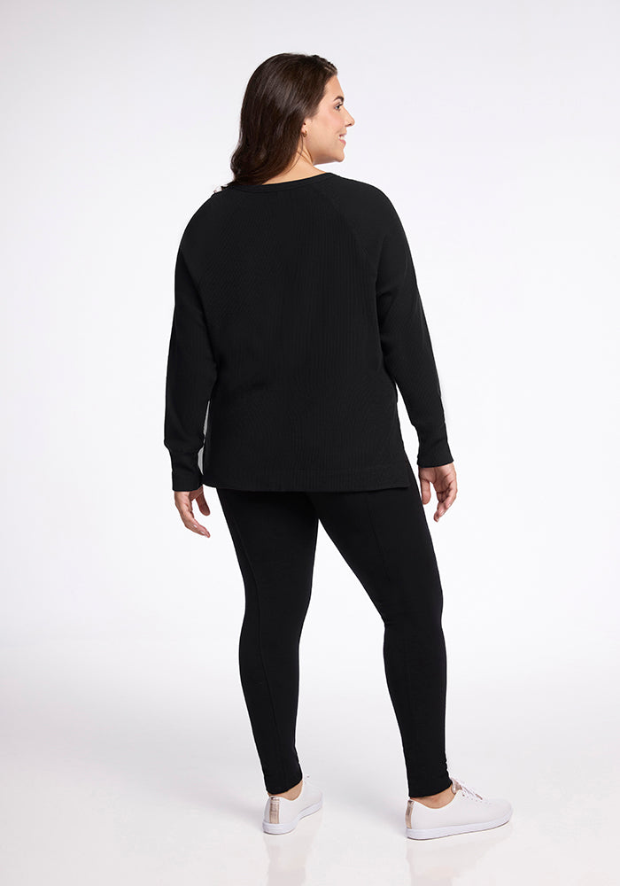 A woman is photographed from the back, standing against a plain white background. She is wearing the Woolx Ainsley Sweater in black, paired with black leggings and white sneakers. Her dark hair falls past her shoulders.