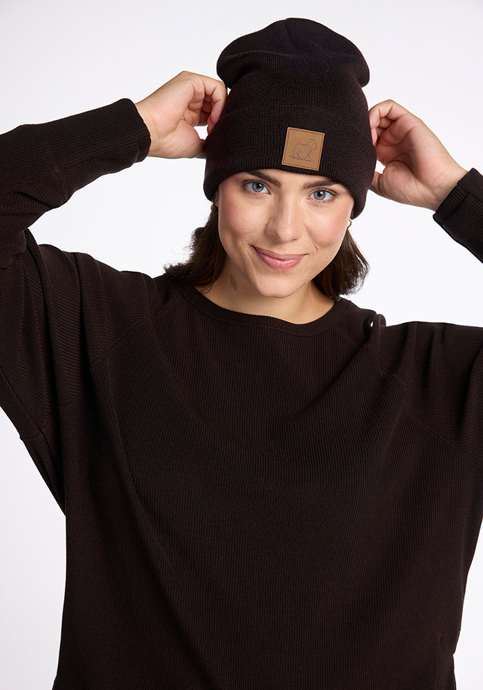 A person with brown hair is wearing a black Woolx Baylor Beanie and a matching black sweater. They are looking at the camera with a slight smile, adjusting the winter hat with both hands against a plain white background.