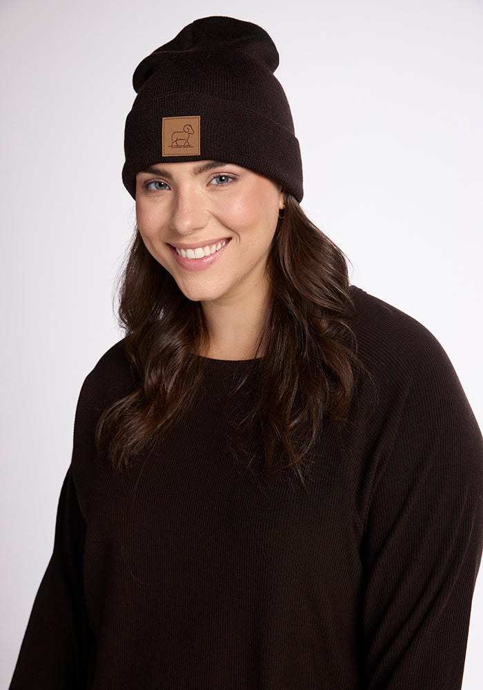 A person with long dark hair wearing a black Woolx Baylor Beanie, which features a brown patch on the front, is smiling at the camera. They are also dressed in a black sweater. The background is plain white.