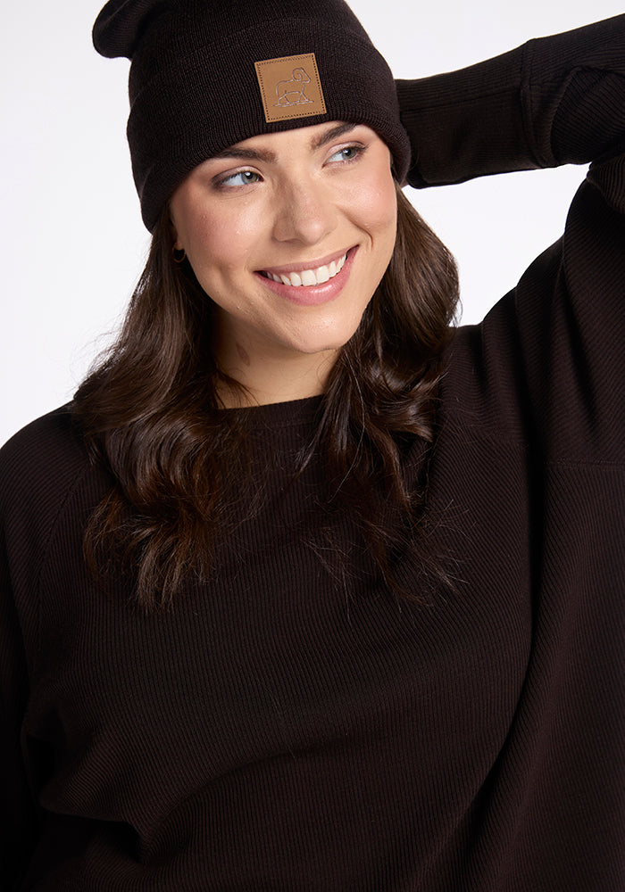 A person with long, dark hair is smiling while wearing a Woolx Baylor Beanie - French Roast and a matching black sweater. The person has one hand raised, touching the back of their head. The warm hat features a small brown leather patch on the front.