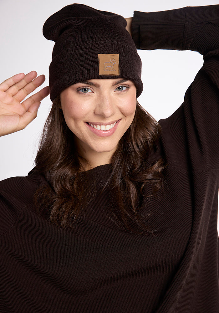 A smiling woman with long, dark hair wears a dark brown Baylor Beanie from Woolx, which features a patch on the front, along with a matching dark brown sweater. She has one hand adjusting the cold weather hat and the other raised to the side of her face against a plain white background.