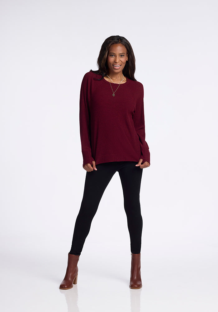 A woman stands smiling against a plain white background. She is wearing a Woolx Ainsley Sweater in Cranberry Melange, black leggings, and brown ankle boots. She also has a necklace and her hands are relaxed by her sides.