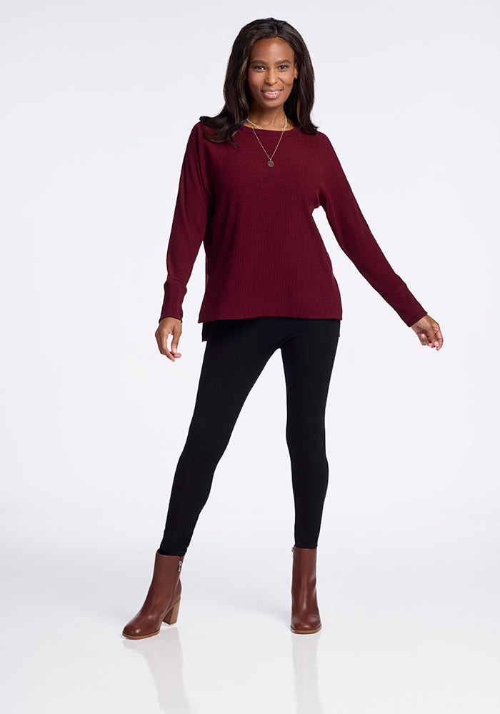 A woman with long dark hair wears a Woolx Ainsley Sweater in Cranberry Melange, black leggings, and brown ankle boots. She is smiling, standing with her left hand on her hip and her right hand relaxed by her side. The background is plain white.