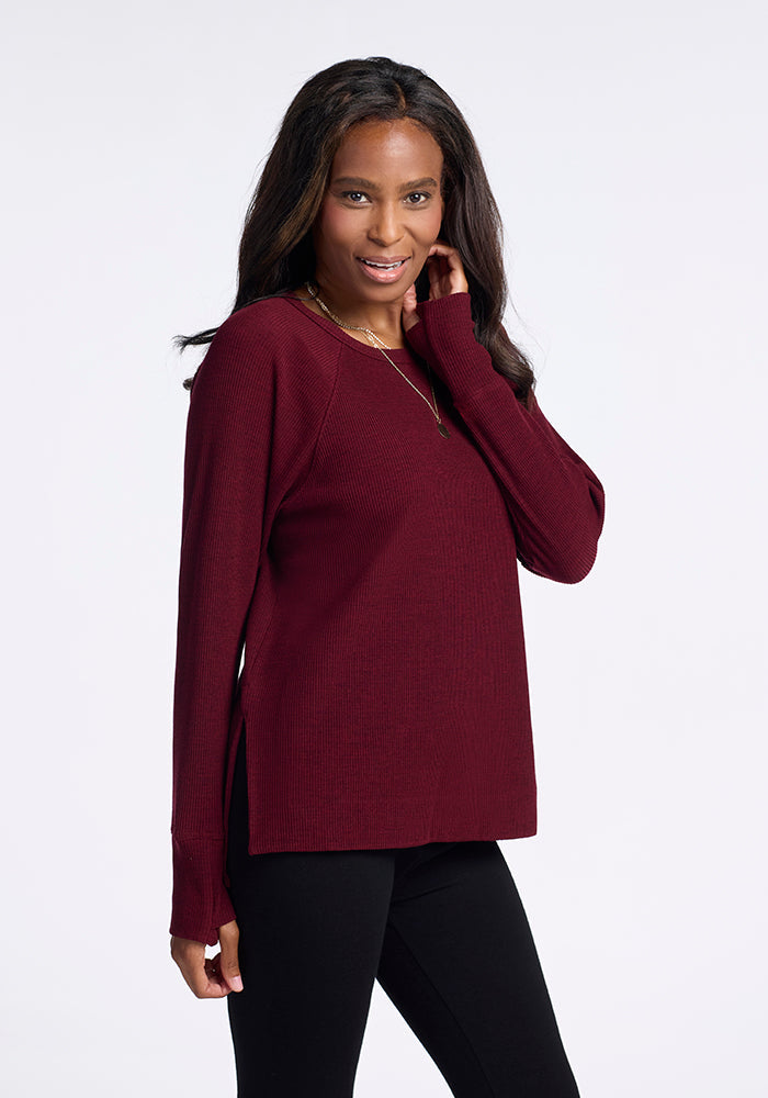 A woman is standing against a plain background, smiling and looking slightly to the side. She is wearing the Ainsley Sweater in Cranberry Melange by Woolx with black pants. She has long, dark hair. One hand is touching her sweater near her face, and the other is by her side. 