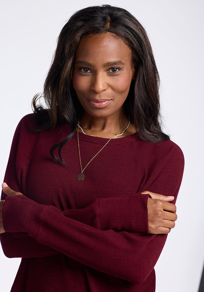 A woman with long, dark hair in a maroon sweater is standing against a plain background. She is smiling and crossing her arms while wearing the Ainsley Sweater - Cranberry Melange by Woolx and a delicate gold necklace with a round pendant.