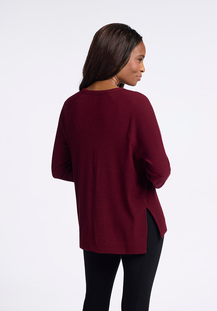 A woman with long hair is standing with her back turned to the camera, wearing the Ainsley Sweater in Cranberry Melange by Woolx, paired with black pants. The background is plain white.