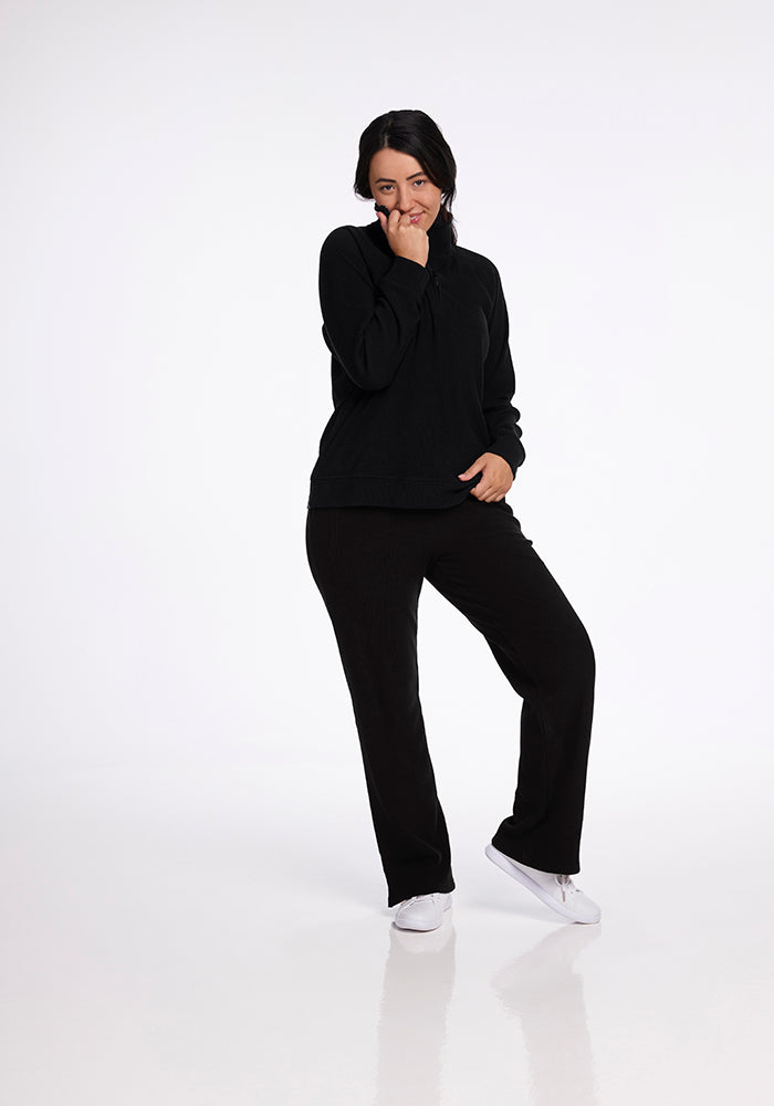 A person wearing the Woolx Quinn Ribbed 1/4 Zip in black and white shoes smiles while standing on one leg. The background is a plain, light color, enhancing the minimalistic look that highlights the elegance of the Woolx top.