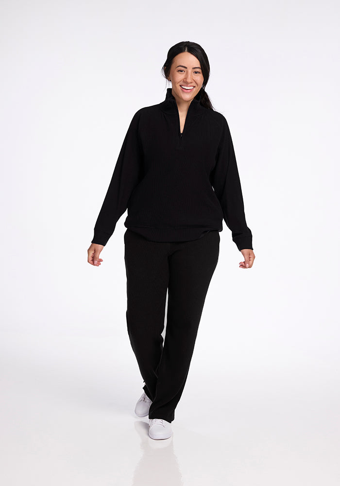 A person with long dark hair is smiling and walking towards the camera. They are wearing the Woolx Quinn 1/4 Zip in black along with matching black pants, paired with white sneakers. The background is white and plain.