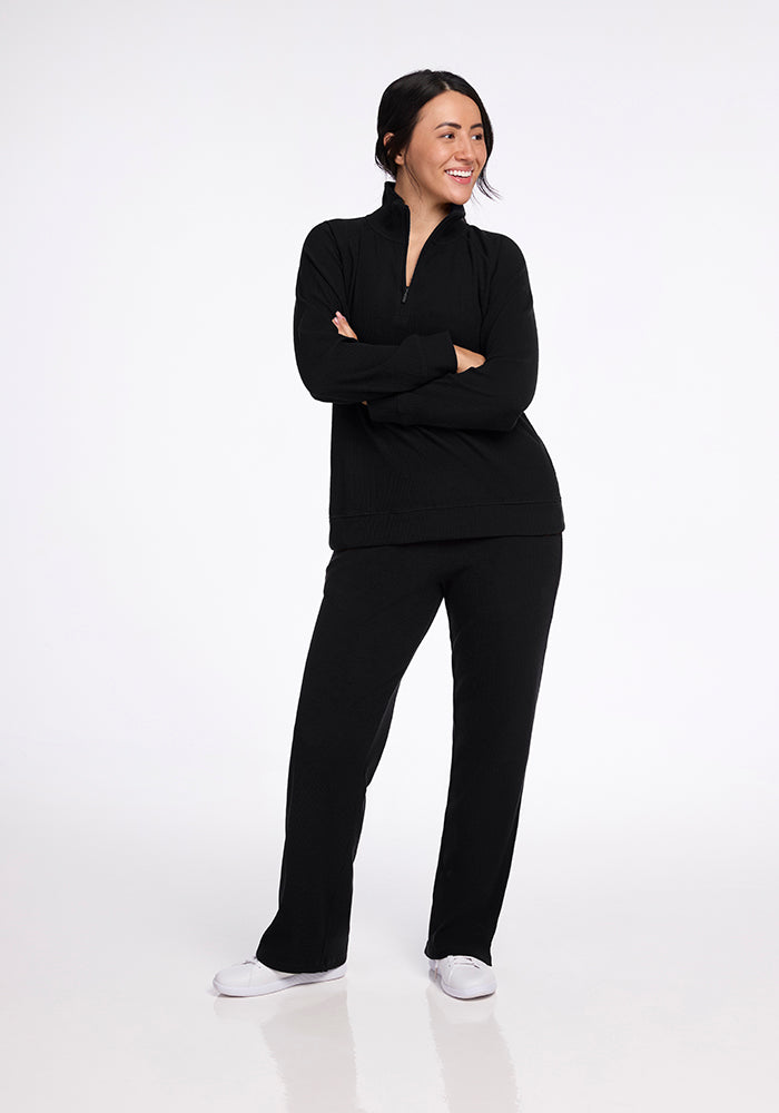 A person stands confidently in a Woolx Quinn Ribbed 1/4 Zip Top in black, paired with matching pants and accented by clean white shoes. They smile warmly with their arms crossed against a plain white background.