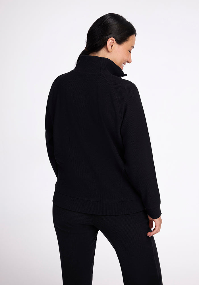 A person with long dark hair stands with their back to the camera, wearing the Quinn Ribbed 1/4 Zip sweater in black by Woolx along with matching pants. The background is plain white.