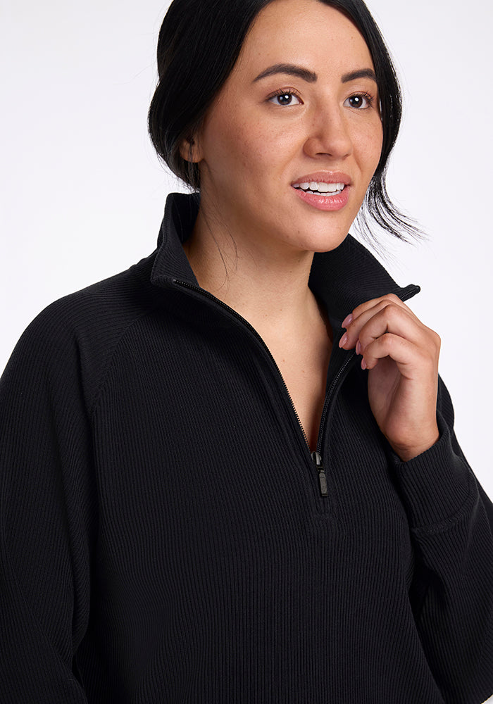 A person with long dark hair is wearing the Quinn Ribbed 1/4 Zip - Black sweater by Woolx, made from Merino wool. They are gazing slightly upward and smiling softly, with a hand gently touching the collar. The background is plain white.