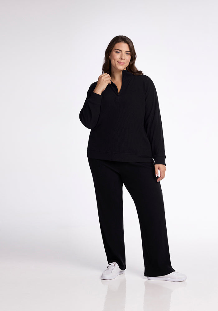 A person stands against a plain white background, wearing the Quinn Ribbed 1/4 Zip in black from Woolx, along with black pants and white sneakers. They are smiling with one hand gently touching their neck.